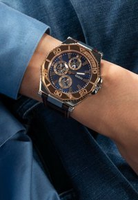 Reloj Guess con esfera azul, caja de acero inoxidable en tonos dorado y plateado, correa de cuero marrón y funciones de cronógrafo.