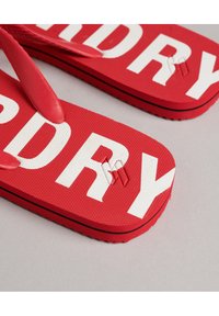 Tongs rouges avec lettrages blancs, dotées d'un semelle texturée, de bandes en caoutchouc et d'un contour noir sur la semelle.