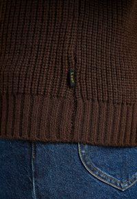 Brun stickad tröja med ribbad nederkant och en liten svart etikett som visar "Lee" i gult. I kombination med blå denimjeans.