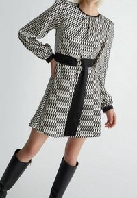 Abito geometrico in bianco e nero con maniche lunghe, cintura nera legata in vita, abbinato a stivali neri al ginocchio.