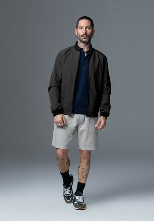 Veste bomber noire avec des poignets côtelés, polo bleu marine, shorts gris clair, et baskets sombres avec des lacets blancs, sur un modèle tatoué.