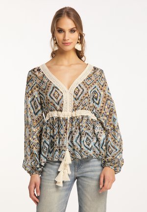 Frau trägt eine farbenfrohe Bluse mit geometrischem Muster, Spitzenbesatz, Quastenband an der Taille und hellblaue Jeans, steht vor weißem Hintergrund.
