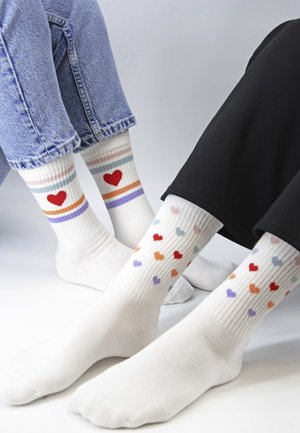 Twee mensen dragen witte sokken met kleurrijke hartjespatronen; de een heeft gestreepte boorden met een rood hart, de ander heeft meerdere kleine hartjes.