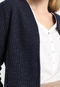 Námořnický modrý texturovan�ý cardigan s tkaným vzorem, zkombinovaný s bílou košilí na knoflíky s malými puntíky a béžovou sukní.