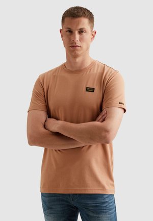 Mladić s kratkom kosom, obučen u bež majicu kratkih rukava s logom na zakrpama, s prekriženim rukama, stoji nasuprot jednobojnoj sivoj pozadini.