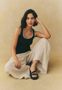 Sort tanktop med dekorativ hvid kant, cremede brede bukser og sorte sandaler. Model sidder, hånd hviler på knæet. Neutral baggrund.