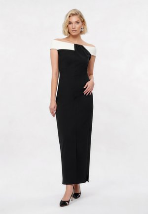 Γυναίκα που φοράει μαύρο φόρεμα χωρίς ώμους με λευκή ταινία στο ντεκολτέ, στέκεται με το ένα χέρι στη μέση και φοράει μαύρες γόβες.