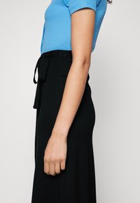 Vue latérale d'une personne portant un haut bleu à manches courtes et une jupe noire taille haute avec une ceinture nouée à la taille.