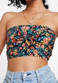 Femme portant un top bandeau floral coloré avec un nœud à l'avant, des colliers empilés en or, des boucles d'oreilles en créole et un jean en denim taille haute bleu clair.