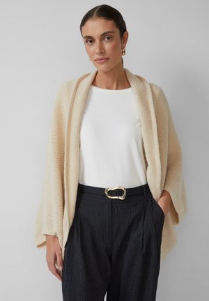 Cardigan en maille couleur crème avec un devant ouvert, associé à un haut blanc et un pantalon à rayures, avec une boucle de ceinture dorée en accent.