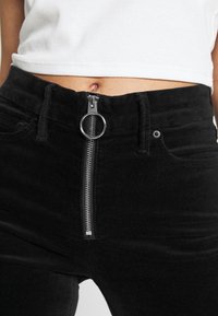 Černé manšestrové kalhoty s předním zipem se stříbrným kruhovým kováním, poutky na pásek a předními kapsami. Hladká textura s přiléhavým střihem.