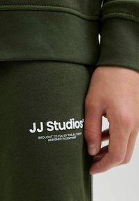 Olīvu zaļš ketšups ar teksturētu virsmu, ar baltu logotipa tekstu "JJ Studios" un etiķetes detaļām kontrastējošā fontā.