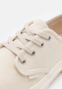 Béžové plátěné tenisky s kulatými stříbrnými očky, plochými tkaničkami a texturovanou podešví. Na boku je logo.