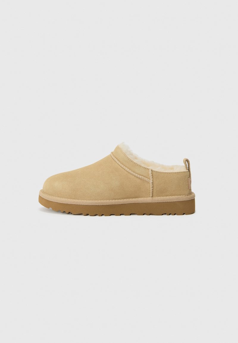 Chausson beige en daim à enfiler avec une semelle épaisse en caoutchouc et une doublure en polaire blanche et duveteuse, conçu pour le confort intérieur et extérieur.