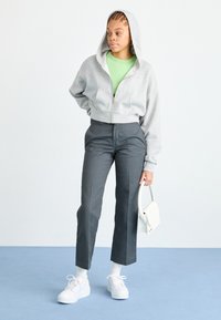 Sweat à capuche gris court avec fermeture éclair devant, t-shirt vert en dessous, pantalon noir taillé, baskets blanches et petit sac à main blanc.