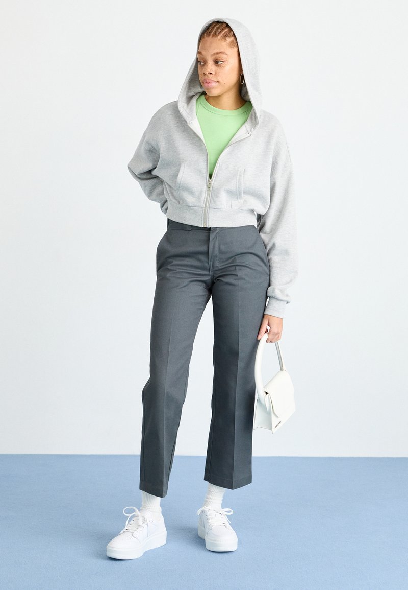Sweat à capuche gris court avec fermeture éclair devant, t-shirt vert en dessous, pantalon noir taillé, baskets blanches et petit sac à main blanc.