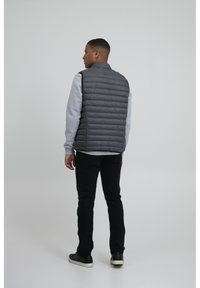 Chaleco acolchado gris sobre un sudadera gris claro, jeans negros y zapatillas negras, visto desde la parte trasera sobre un fondo liso.