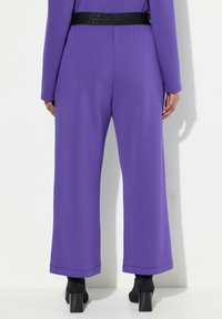 Lila weit geschnittene Hose aus weichem Material mit einem schwarzen elastischen Bund mit Branding, entworfen für Komfort und Bewegungsfreiheit.