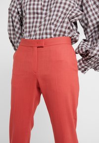 Koraalkleurige broeken met een gladde textuur, met een platte front en een getailleerde pasvorm, gecombineerd met een geruite blouse met lange mouwen.