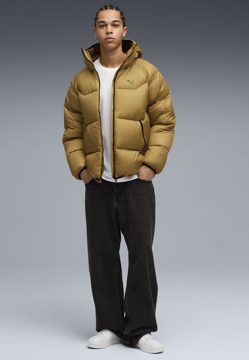 Giacca puffer gialla senape con cappuccio, dotata di zip frontale, polsini elasticizzati e dettagli neri. Abbinata a pantaloni scuri e sneakers bianche.