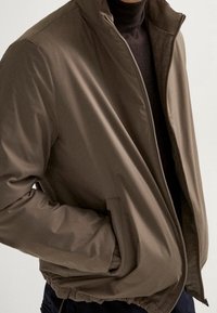Brown, zipped jacket with a high collar, featuring a smooth exterior and side pockets, paired with a dark turtleneck underneath.
