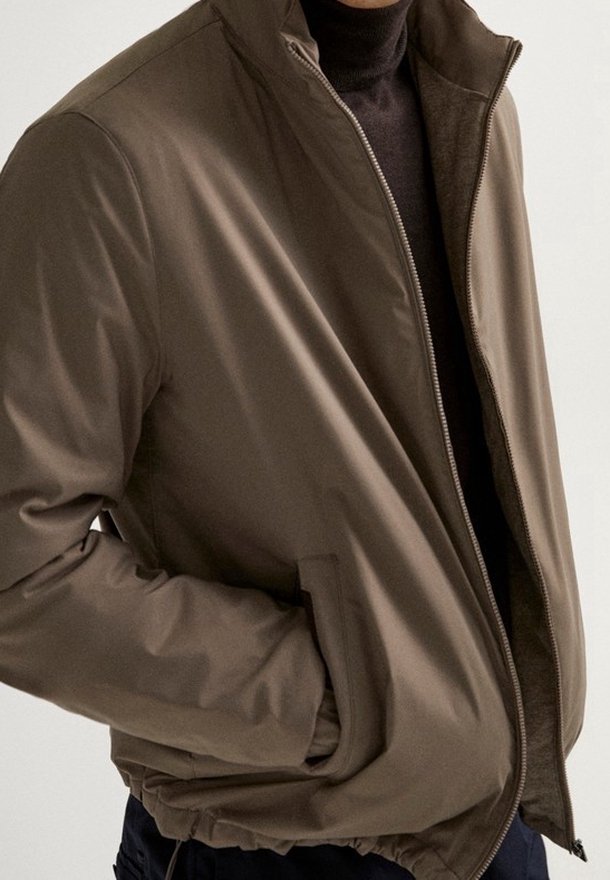 Brown, zipped jacket with a high collar, featuring a smooth exterior and side pockets, paired with a dark turtleneck underneath.