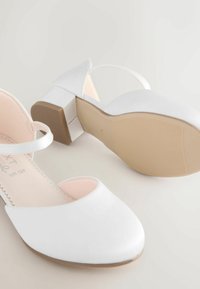 Valkoiset korkokengät, joissa on pyöreä kärki, nilkkahihna ja lohkokorko. Tasainen materiaali, jossa on bežsäkäinen pohja. Minimalistinen muotoilu, ei kuvioita.