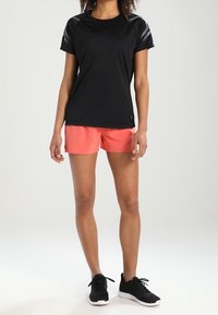 Camiseta deportiva negra con detalles de malla, combinada con pantalones cortos coral brillantes. El modelo lleva zapatillas deportivas negras y está de pie sobre un fondo liso.