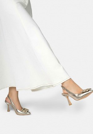 Voeten met metallic slingback hoge hakken met met juwelen versierde gespen, gecombineerd met vloeiende witte brede broekspijpen.