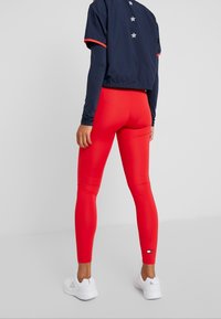 Rote Leggings mit glatter Textur und hohem Bund, kombiniert mit einer marineblauen Cropped-Jacke mit weißen Sternen auf dem Rücken und orangefarbenen Akzenten. Weiße Sneakers.