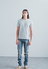 Jovem com cabelo escuro até aos ombros, vestindo uma t-shirt cinza claro, jeans azuis desbotados e sapatos bege, encostado a uma parede simples.