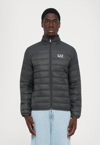 Svart, quilted, glidelåsjakke med EA7 Emporio Armani-logo, båret av en stående mann i lyseblå jeans, vendt mot kameraet foran en ensfarget bakgrunn.