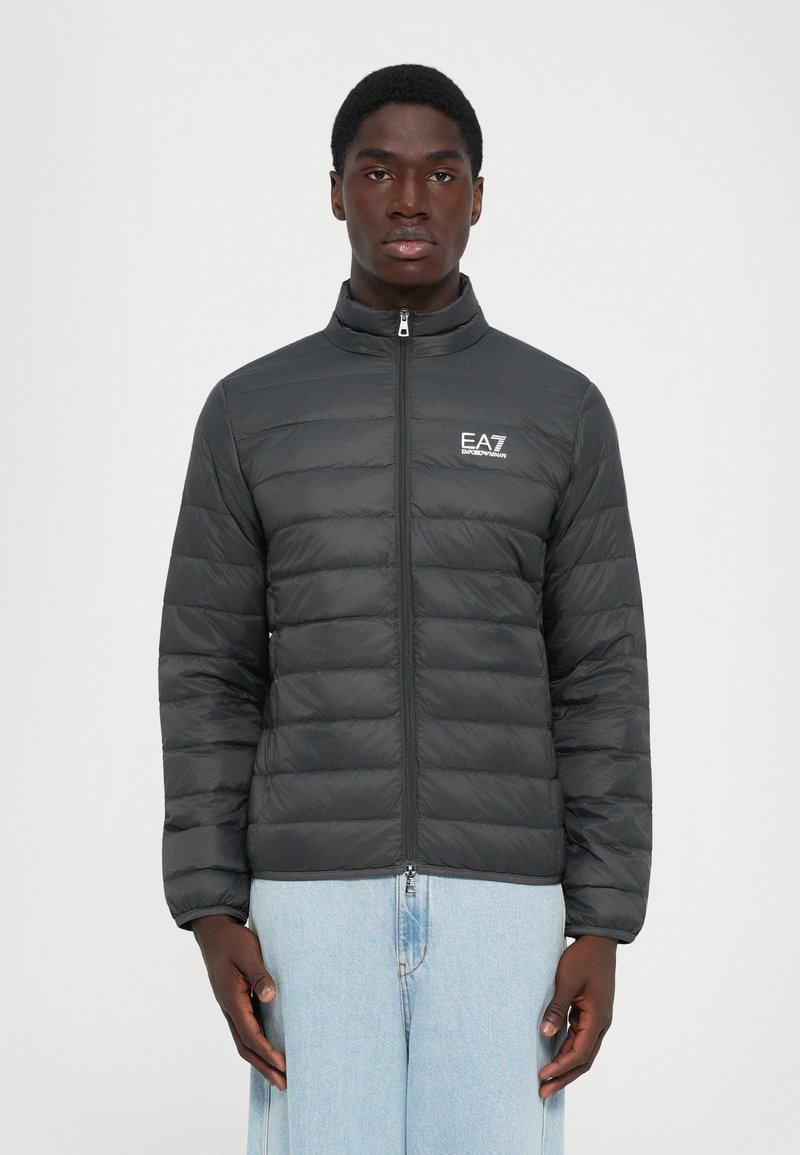Svart, quilted, glidelåsjakke med EA7 Emporio Armani-logo, båret av en stående mann i lyseblå jeans, vendt mot kameraet foran en ensfarget bakgrunn.