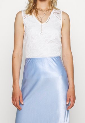 Kobieta w białej, bezrękawnikowej koronkowej bluzce, jasnoniebieskiej satynowej spódnicy i warstwowych złotych naszyjnikach z zawieszką.