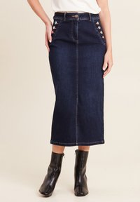 Gonna lunghèzza media in denim scuro con taglio dritto, accentuata da bottoni laterali e cuciture a contrasto, abbinata a stivali neri alla caviglia.