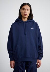 Ναυτικό μπλε φούτερ με κουκούλα φτιαγμένο από μαλακό ύφασμα, με μπροστινή τσέπη. Λευκό λογότυπο Nike στο επάνω αριστερό μέρος. Διαθέτει χαλαρή εφαρμογή και κουκούλα με κορδόνι.