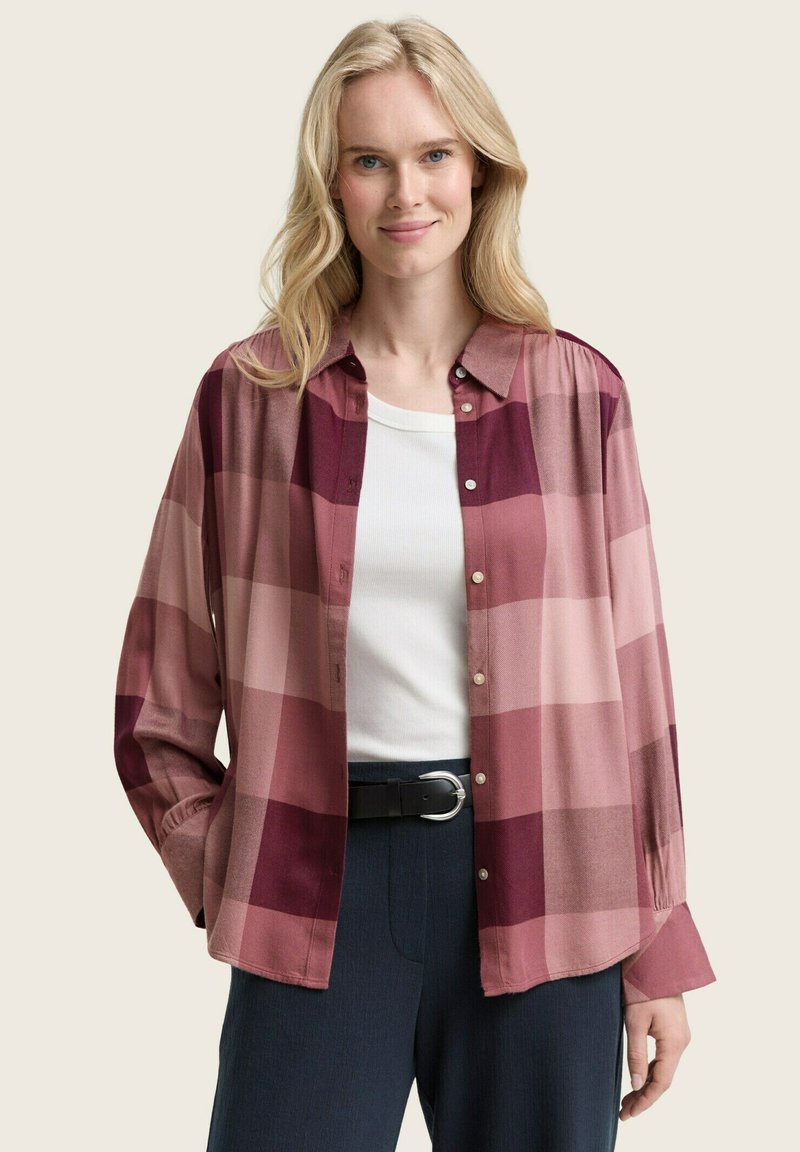 Chemise à boutons en motif tartan rose avec des teintes variées, en tissu doux, à manches longues et avec un col, portée avec un top blanc.