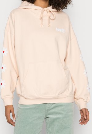 Personne portant un sweat à capuche Levi's rose clair avec des marguerites blanches sur les manches et un pantalon en velours côtelé vert clair.