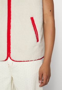 Gilet en polaire de couleur crème, avec une fermeture éclair et des bordures rouges, doté d'une poche latérale et d'une surface douce et texturée.