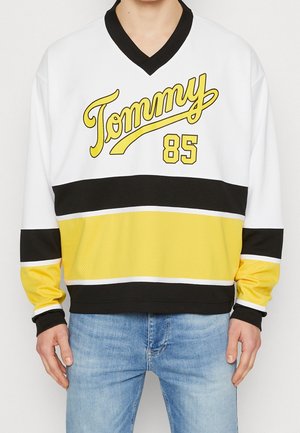 Sweat-shirt blanc, jaune et noir avec le texte "Tommy 85" porté par une personne en jean bleu, debout devant un fond clair uni.