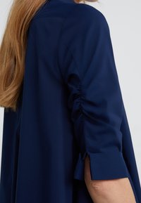 Mörkblå blus med samlade ärmar och uppvikta manschetter. Mjukt tyg med en slät struktur som visar en modern och strukturerad design.