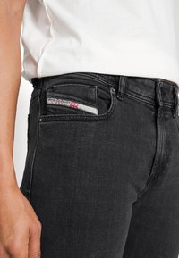 Vaqueros de mezclilla negros con un ajuste ceñido, diseño de cinco bolsillos y una etiqueta de marca visible en el bolsillo superior derecho. Textura suave, detalles mínimos.