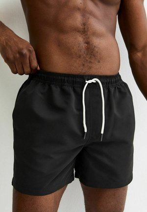 Uomo che sistema pantaloncini da bagno neri con coulisse bianca, mostrando il torso nudo e gambe muscolose su sfondo neutro.