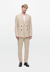 Beige, doppelt geknöpfter Anzugjacke mit schwarzen Knöpfen, kombiniert mit passender Hose. Darunter ein weißes Hemd und schwarze Schuhe. Glatte Textur.