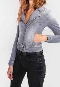Džínová bunda v barvě světle šedé s cropped designem, zipovým zavíráním, špičatým límcem a dlouhými rukávy; má boční kapsu a knoflíkové detaily.