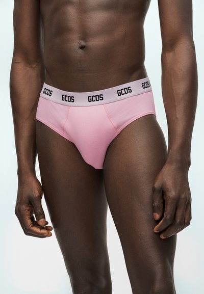 Άνδρας μοντέλο που φοράει ροζ εσώρουχα με ανοιχτόχρωμη μέση που φέρει το λογότυπο "GCDS", στέκεται μπροστά σε ένα απλό φωτεινό φόντο.