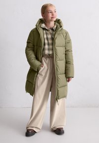 Long, puffy green parka jacket with a hood, featuring a quilted design. Worn over a plaid shirt and beige wide-leg trousers.