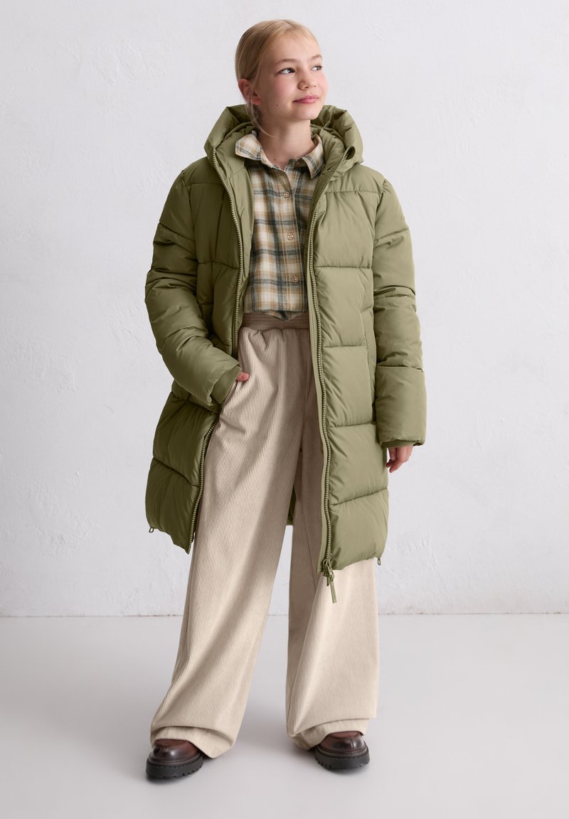 Long, puffy green parka jacket with a hood, featuring a quilted design. Worn over a plaid shirt and beige wide-leg trousers.