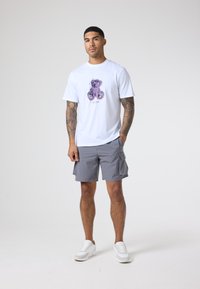 Hvit t-skjorte med et lilla bjørnegrafikk og grå cargo-shorts. Fottøyet er hvite sneakers. Bakgrunnen er ensfarget og nøytral.