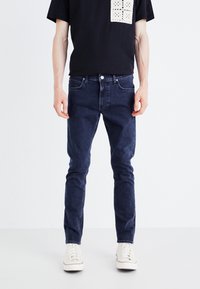Jeans bleu foncé au design slim, avec une taille standard et un style classique à cinq poches. Portés avec un t-shirt noir.